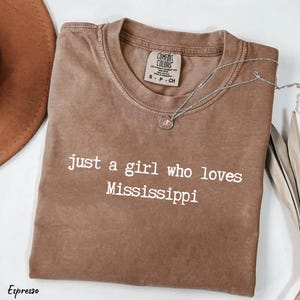 May include: Espresso-colored t-shirt with the text "just a girl who loves Mississippi" in white. A silver necklace with a pendant rests on the shirt. The shirt is folded, with a brown hat visible in the background.