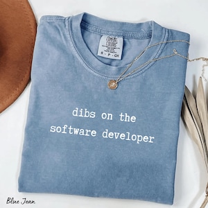 May include: A blue jean coloured t-shirt with the text "dibs on the software developer" in white. A gold necklace with a pendant is draped on the shirt. The shirt is folded and has a tag from Comfort Colors.