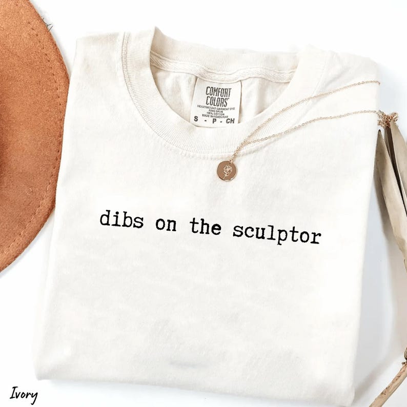 Op de afbeelding: Ivoor kleurig t-shirt met de tekst "dibs on the sculptor" in het zwart gedrukt. Een goudkleurige ketting met een hanger ligt over het shirt. Een bruine hoed is gedeeltelijk zichtbaar in de linkerbovenhoek.