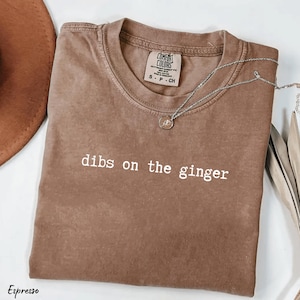 May include: Espresso-coloured t-shirt with the phrase "dibs on the ginger" in white lettering. The shirt has a crew neck and short sleeves. A silver necklace with a pendant is draped across the shirt. A brown hat is partially visible.