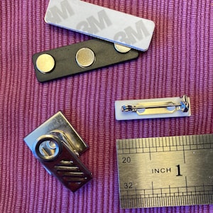 May include: A collection of name tag components, including a white rectangular label with 3M adhesive, a black magnetic bar with two magnets, a white pin-back label, and a silver metal clip. A ruler shows the length of the items in inches.