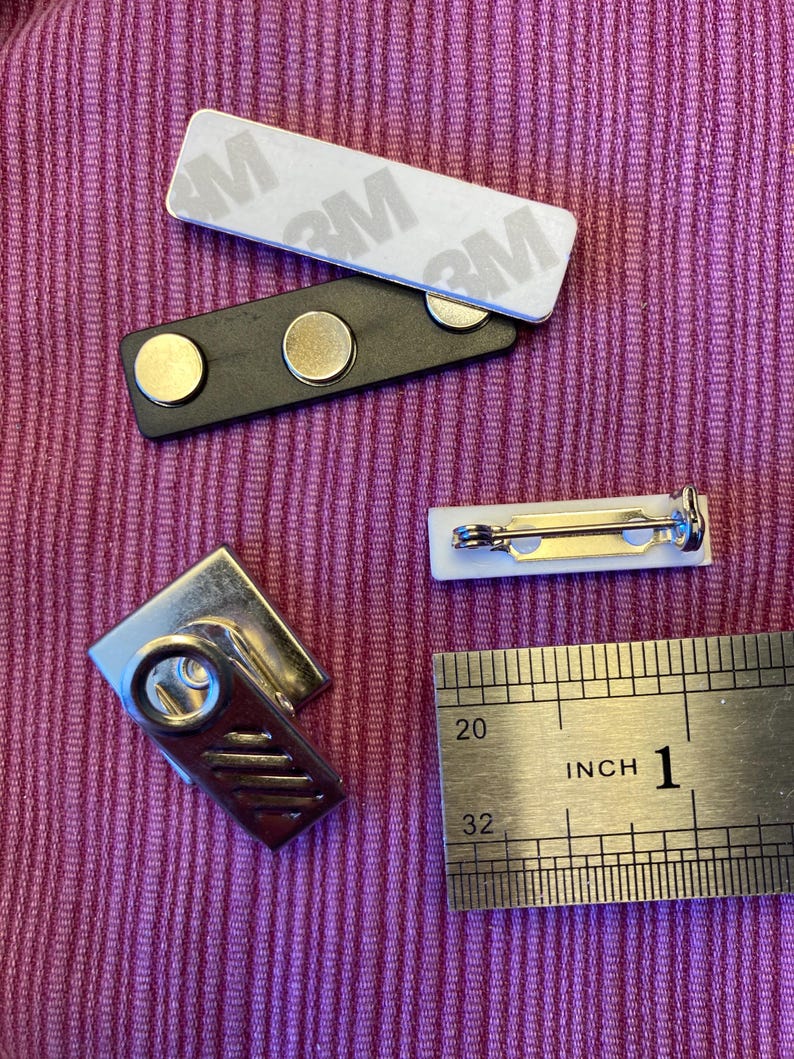 May include: A collection of name tag components, including a white rectangular label with 3M adhesive, a black magnetic bar with two magnets, a white pin-back label, and a silver metal clip. A ruler shows the length of the items in inches.