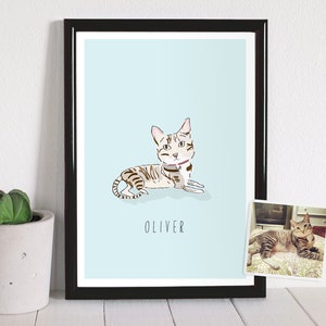 May include: A framed art print featuring a stylized illustration of a cat named Oliver, lying down on a light blue background. The cat is depicted with brown and gray stripes and a pink collar. A small photo of the cat is in the lower right corner.
