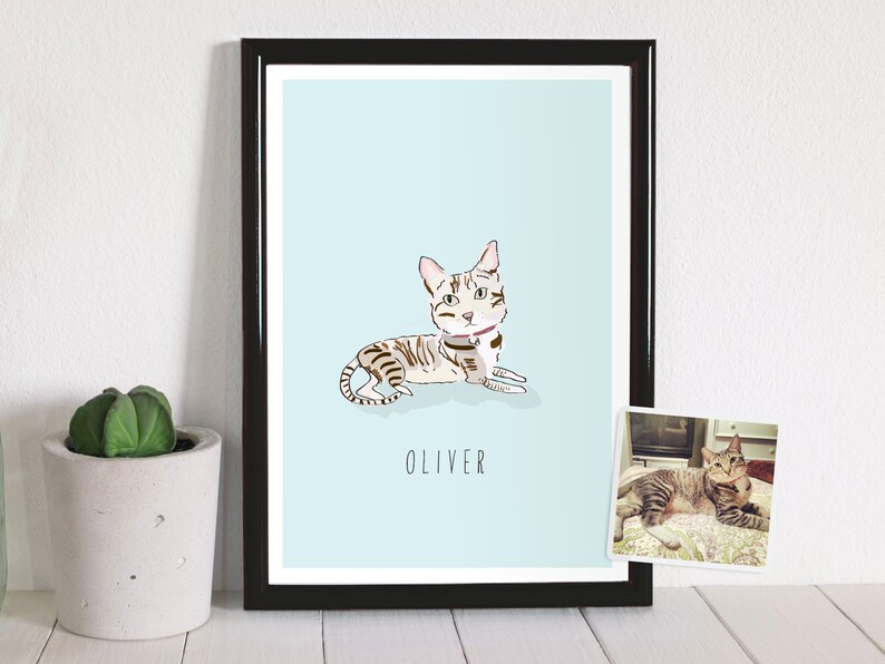 May include: A framed art print featuring a stylized illustration of a cat named Oliver, lying down on a light blue background. The cat is depicted with brown and gray stripes and a pink collar. A small photo of the cat is in the lower right corner.