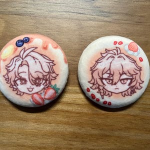 May include: Two round, white and pink decorative pins with anime-style character illustrations. One pin features strawberries and blueberries, the other a strawberry. The pins have a cartoonish design with a light pink and white color scheme.