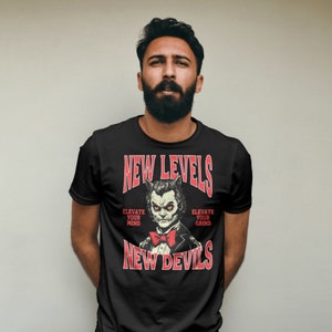 Peut inclure: T-shirt noir avec un graphique rouge d'un loup de dessin animé portant un nœud papillon. Le graphique indique "New Levels New Devils" avec le texte "Elevate Your Mind" et "Elevate Your Grind" de chaque côté du loup.