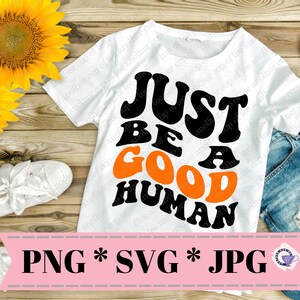 May include: White t-shirt with the text "Just Be A Good Human" in black and orange retro font. The shirt is displayed on a wooden surface with a sunflower and a pair of white shoes.