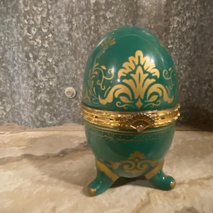 May include: A green porcelain egg-shaped trinket box with gold accents and a floral design. The box has three legs and a hinged lid.
