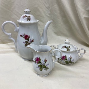 May include: A white porcelain tea set featuring a teapot, creamer, and sugar bowl. Each piece is decorated with a floral rose design in shades of pink and green, with gold trim accents. The set is displayed on a white surface.