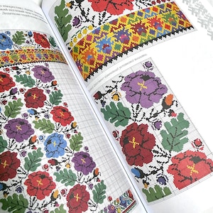 May include: Open book with pages displaying colorful cross-stitch patterns. The designs feature vibrant red, purple, and blue flowers with green leaves. The patterns are set against a grid background.