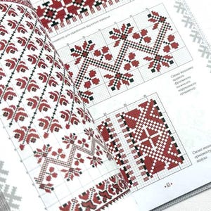 May include: Open book displaying cross-stitch patterns in red, black, and white. The designs feature geometric and floral motifs, with grid lines for guidance. Text in the image is in a foreign language.