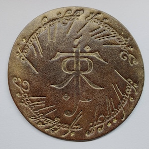May include: A round, brown leather coaster with a gold inscription around the edge. The inscription reads "One Ring to rule them all, One Ring to find them, One Ring to bring them all and in the darkness bind them." In the center of the coaster is a gold inscription of the word "The" in the Elvish language.