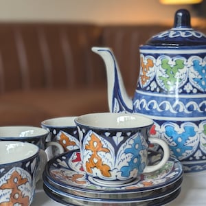 May include: A blue and white ceramic tea set with a teapot, cups, and saucers. The set features a decorative leaf pattern in orange, green, and blue. The cups and saucers are stacked, and the teapot is visible in the background.