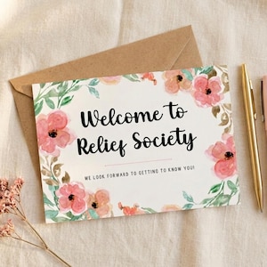 May include: A rectangular greeting card with a white background featuring watercolour-style pink flowers and green leaves. The text reads Welcome to Relief Society and WE LOOK FORWARD TO GETTING TO KNOW YOU! A gold pen and a brown envelope are visible.
