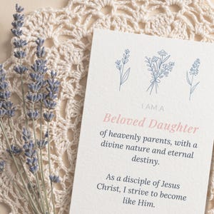 Może przedstawiać: Karta z napisem "I AM A Beloved Daughter" i ilustracjami kwiatowymi. Karta leży na kremowej, szydełkowej serwetce, z gałązkami lawendy po lewej stronie. Tekst na karcie kontynuuje przesłanie o boskiej naturze i przeznaczeniu.