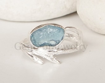 May include: A silver ring with a light blue, raw-cut gemstone. The ring's band has a textured, organic design, with a branch-like element extending from the side. The gemstone is set in a silver bezel.