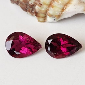 May include: Two pear-shaped gemstones, each a deep, rich red color. The gemstones are set against a white background.