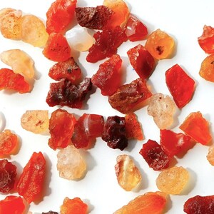 Puede incluir: Una colección de pequeñas piedras preciosas ásperas de color rojo, naranja y blanco. Las piedras están esparcidas sobre una superficie blanca.