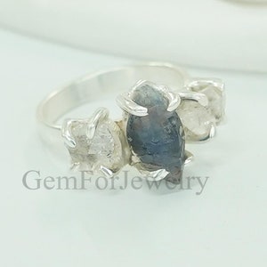 Op de afbeelding: Een zilveren ring met drie ruwe edelstenen. De middelste steen is een donkerblauwe saffier, en de twee zijstenen zijn helder kwarts. De ring is gezet op een eenvoudige band.