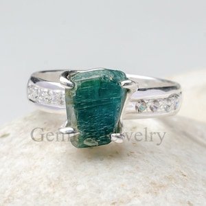 Può includere: Un anello d'argento con una pietra preziosa verde e una fascia di piccole pietre chiare.