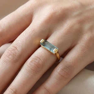 May include: A gold ring featuring a rectangular, light blue gemstone. The ring is set on a slender gold band. The gemstone is a light blue color. The ring is shown on a hand.