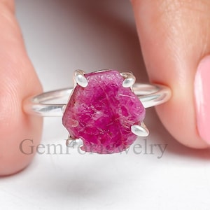 May include: A silver ring featuring a large, raw, pink ruby gemstone. The ruby is set in a simple, claw setting.