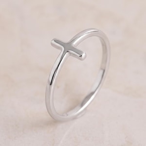 May include: A polished silver-toned ring featuring a small, smooth cross emblem integrated into the thin, circular band. The metal surface is reflective and uniform, showcasing a simple, continuous design with no additional stones or engravings.