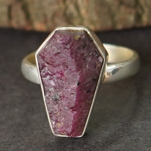 May include: A silver ring with a large, faceted, pink ruby stone shaped like a coffin.