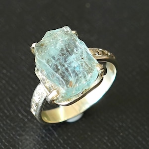May include: A silver ring with a large, light blue aquamarine gemstone. The gemstone is rough and has a natural, unpolished look.