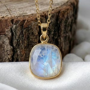 Peut inclure: Un collier doré avec un pendentif en pierre de lune carré et irisé. Le pendentif est serti dans une lunette dorée et est suspendu à une fine chaîne dorée. La pierre de lune affiche des teintes bleues et blanches. Le collier est sur un tissu blanc.