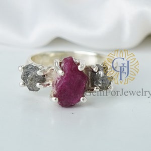 May include: A silver ring with a large, rough-cut pink ruby gemstone and two smaller, rough-cut gray gemstones. The ring is set in a simple band.