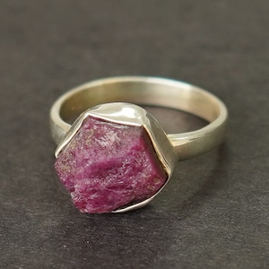 Può includere: Un anello d'argento con una grande pietra preziosa di rubino grezzo incastonata al centro. Il rubino è di colore rosa intenso e ha una texture ruvida e naturale.