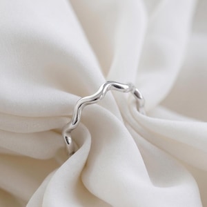 Puede incluir: Un anillo plateado con un diseño ondulado y orgánico. La superficie pulida del anillo refleja la luz, sobre un fondo de tela blanca. La forma inusual del anillo le da un aspecto moderno, adecuado para el uso diario.