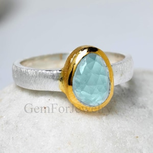 Pode incluir: Um anel de prata com uma moldura dourada que segura uma pedra preciosa facetada azul clara. O anel está sobre uma superfície branca e texturizada. A gema tem uma forma irregular.