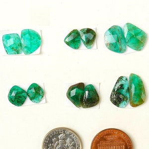 Könnte beinhalten: Eine Auswahl an Smaragd-Edelsteinen in verschiedenen Formen, präsentiert auf kleinen weißen Karten. Die Edelsteine sind von leuchtend grüner Farbe und haben facettierte Oberflächen. Eine amerikanische Dime und ein Penny sind zum Größenvergleich enthalten.