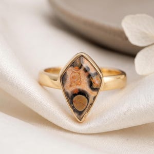 May include: A gold ring featuring a kite-shaped stone with a complex pattern of tan, brown, and black circular inclusions. The polished gold band holds the stone in a smooth bezel setting, creating a distinct geometric silhouette on the finger.