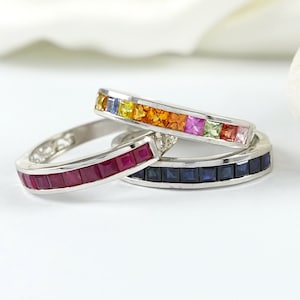 May include: Three eternity bands with a rainbow of gemstones. The top band features orange, yellow, and red gemstones. The middle band features red gemstones. The bottom band features blue gemstones.