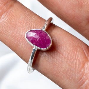 May include: A silver ring featuring a vibrant, oval-shaped ruby gemstone. The ruby has a deep pinkish-red hue and is set in a simple, elegant band. The ring is shown on a finger against a soft, white background.