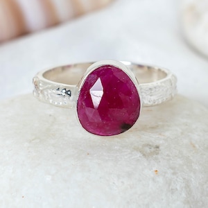 May include: A silver ring with a large, oval-cut ruby gemstone. The ruby is a vibrant, deep red hue. The ring's band has a textured, etched pattern. The ring is presented on a white stone, with other stones softly blurred in the background.