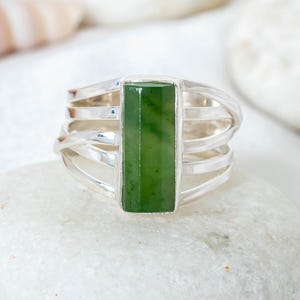 Könnte beinhalten: Ein Silberring mit einem rechteckigen, leuchtend grünen Jadestein. Der Ring hat ein Mehrband-Design mit vier parallelen Bändern auf jeder Seite des Jades. Der Ring wird auf einem weißen Stein präsentiert.