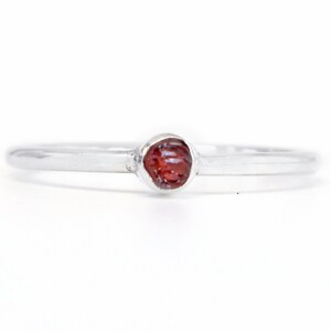 May include: A simple silver band ring with a small, red, raw gemstone set in the center.