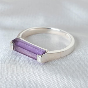 May include: A silver ring featuring a rectangular, light purple gemstone. The gemstone is set in a simple, modern design. The ring is displayed on a white, textured surface, highlighting the ring's polished metal and the gemstone's color.