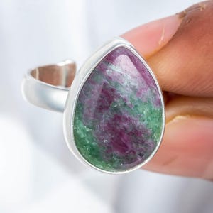 May include: A silver ring featuring a large, teardrop-shaped gemstone. The gemstone displays a mix of deep red, green, and white hues. The ring is held by a person's fingers, showcasing the polished silver band and the stone's unique color pattern.