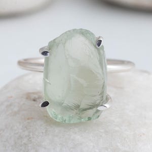 May include: A silver ring featuring a pale green, raw-cut gemstone. The stone is held by four silver prongs. The ring is displayed on a white, textured surface. The gemstone has a unique, irregular shape.