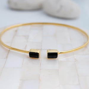 May include: A gold-colored bangle bracelet with two black rectangular stones at the opening. The bracelet is displayed on a white, textured surface. The text "GemForJewelry" is visible on the bracelet.