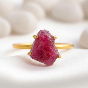 May include: A gold ring featuring a raw, uncut ruby gemstone. The ruby is a deep, vibrant red color and is held in place by four small gold prongs. The ring band is a simple, polished gold. The ring is set against a white background.
