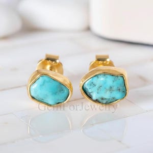 Könnte beinhalten: Ein Paar goldfarbene Ohrstecker mit türkisfarbenen Steineinsätzen. Die Ohrringe haben ein schlichtes, elegantes Design, wobei die Türkissteine in einer goldenen Lünette gefasst sind. Die Ohrringe werden auf einer weißen, reflektierenden Oberfläche präsentiert.