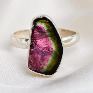 K&ouml;nnte beinhalten: Ein Silberring mit einem gro&szlig;en, unregelm&auml;&szlig;ig geformten Wassermelonen-Turmalin-Edelstein. Der Edelstein zeigt eine lebendige Mischung aus rosa, gr&uuml;n und schwarzen Farben. Der Ring ist vor einem wei&szlig;en, seidigen Hintergrund platziert.