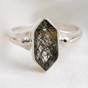 May include: A silver ring with a faceted, elongated gemstone containing black and gold inclusions. The ring's band splits near the stone, with small silver spheres either side of the gem. The ring is set against a white, textured background.
