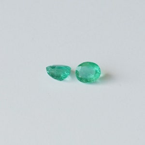May include: Two emerald green gemstones, one slightly larger than the other, are displayed on a white surface.